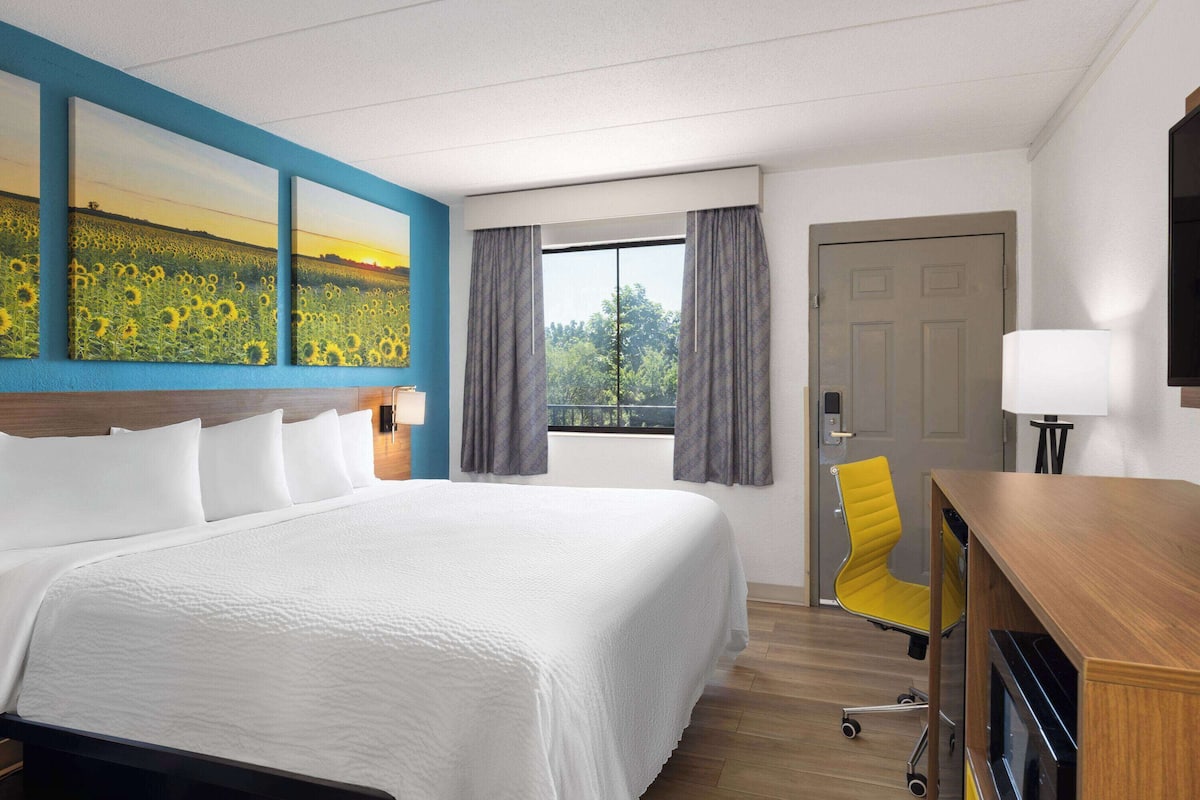 Habitación de hotel con una cama grande, un escritorio con silla, una ventana con cortinas y un cuadro en la pared.