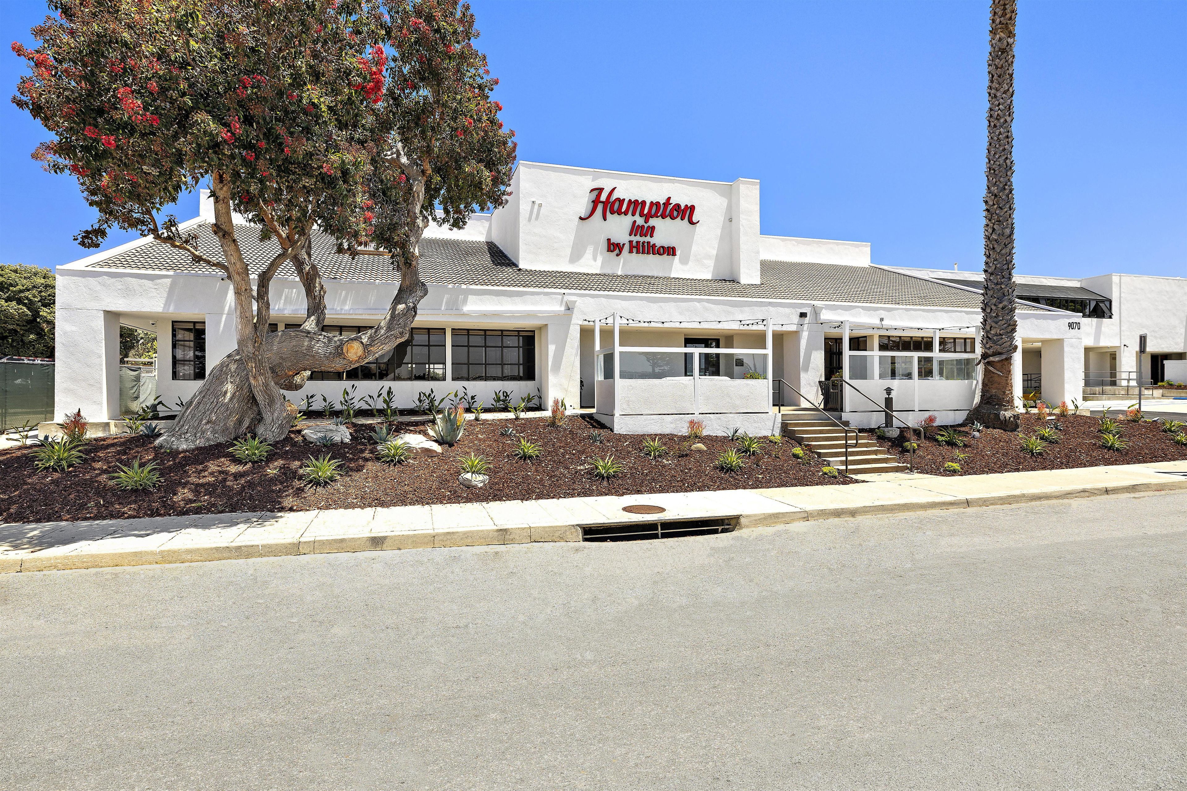 A Hampton Inn by Hilton with a white facade, a tiled roof, and a landscaped front yard.