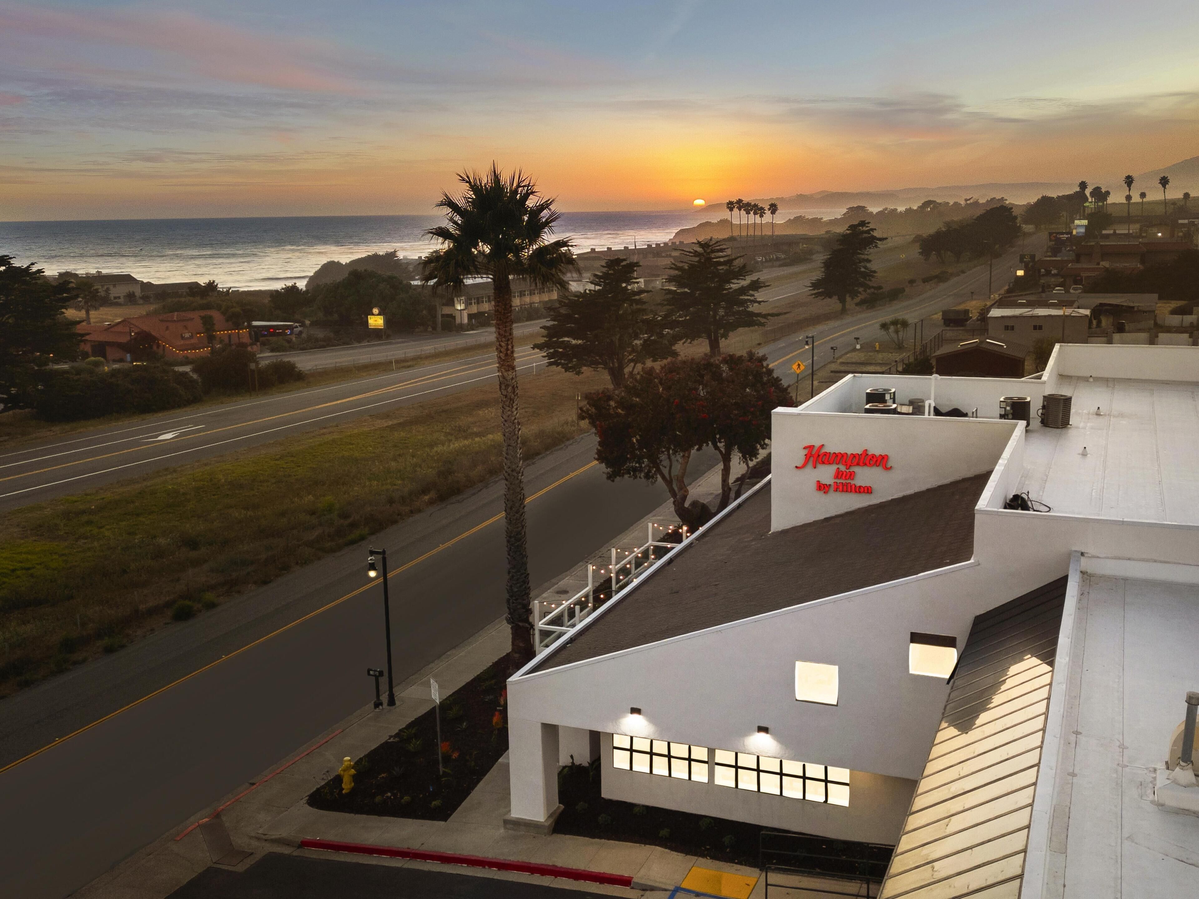 Hotel com vista para o pôr do sol sobre o oceano, uma estrada em primeiro plano e céu claro.