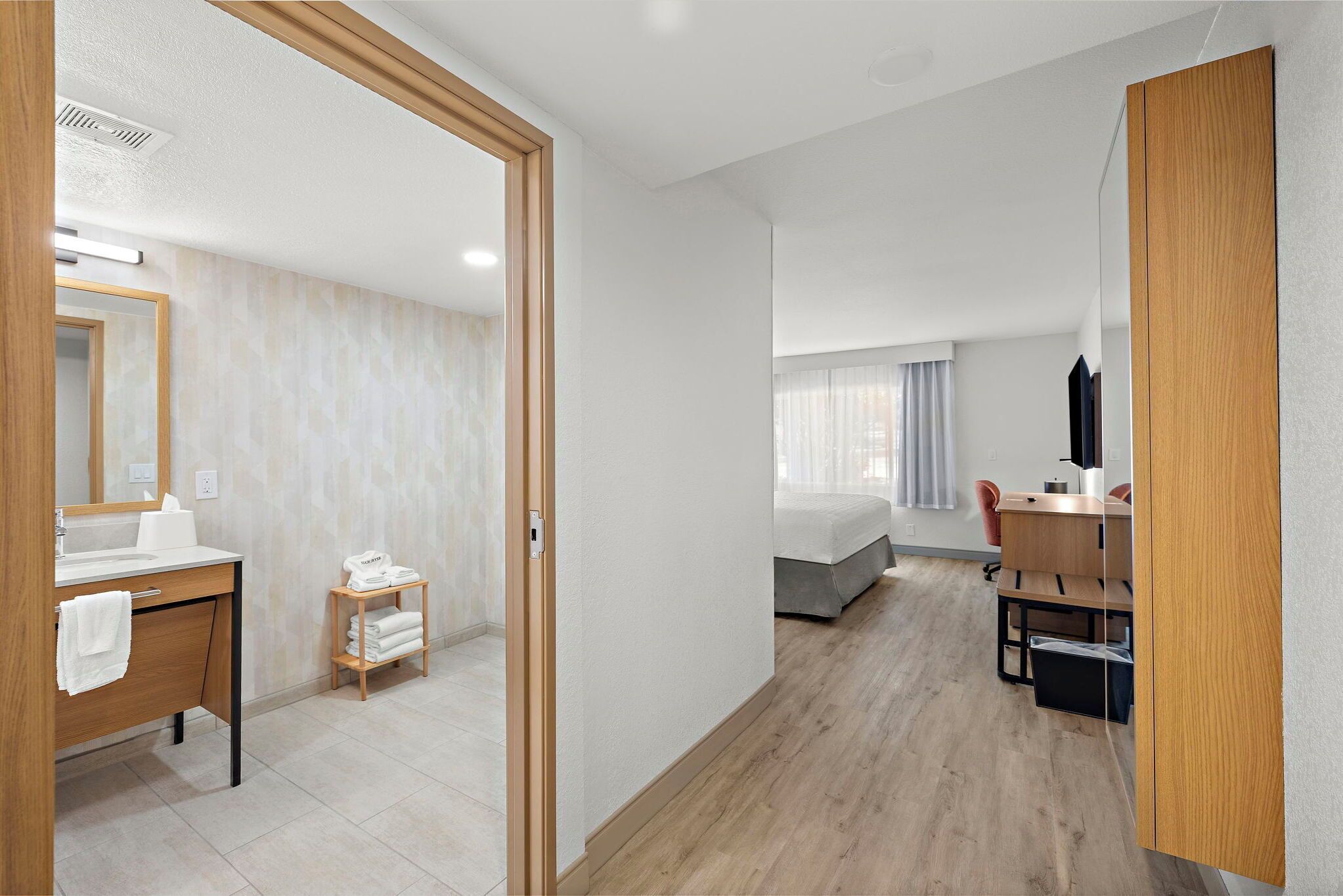 A hotel room with a wooden floor, a bed, a desk with a chair, a mirror, and a window with curtains.