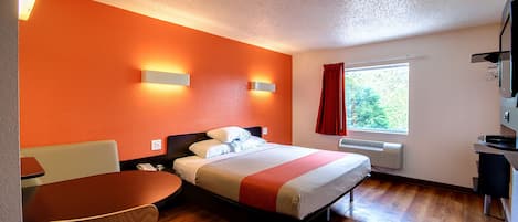 A hotel room with a bed, a desk, and a chair. There is a window with a view of trees.
