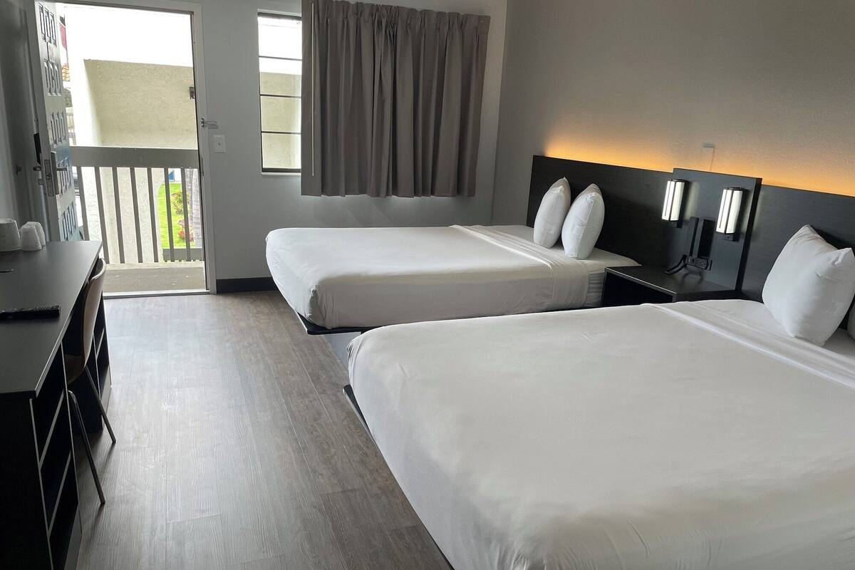 Une chambre d’hôtel avec deux lits simples, un bureau avec une chaise, une table de chevet et une fenêtre avec des rideaux.