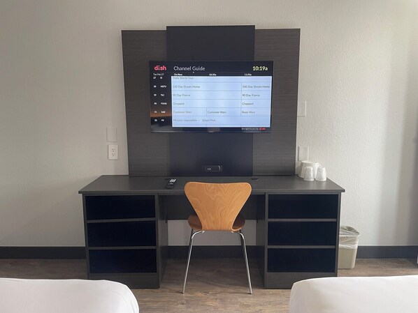 Habitación de hotel con escritorio, televisor mostrando una guía de canales, una silla y dos camas.