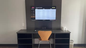 Habitación de hotel con escritorio, televisor mostrando una guía de canales, silla de madera y un cesto de basura.