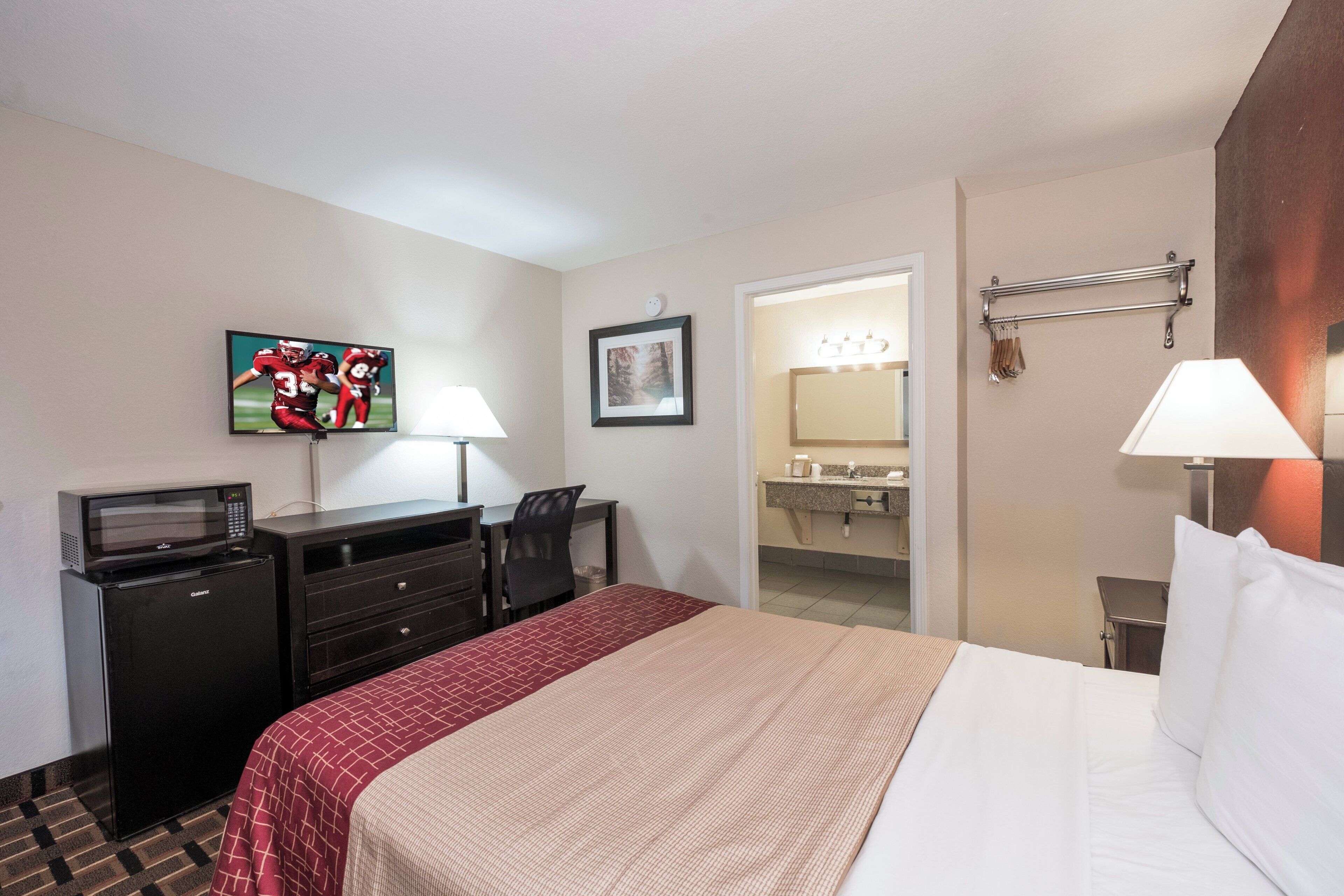 A hotel room with a bed, a desk with a computer, a TV, a mirror, and a bathroom visible through an open door.