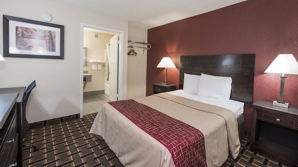A hotel room with a bed, two bedside lamps, a desk, and a chair. There is a framed picture on the wall and a door leading to a bathroom.
