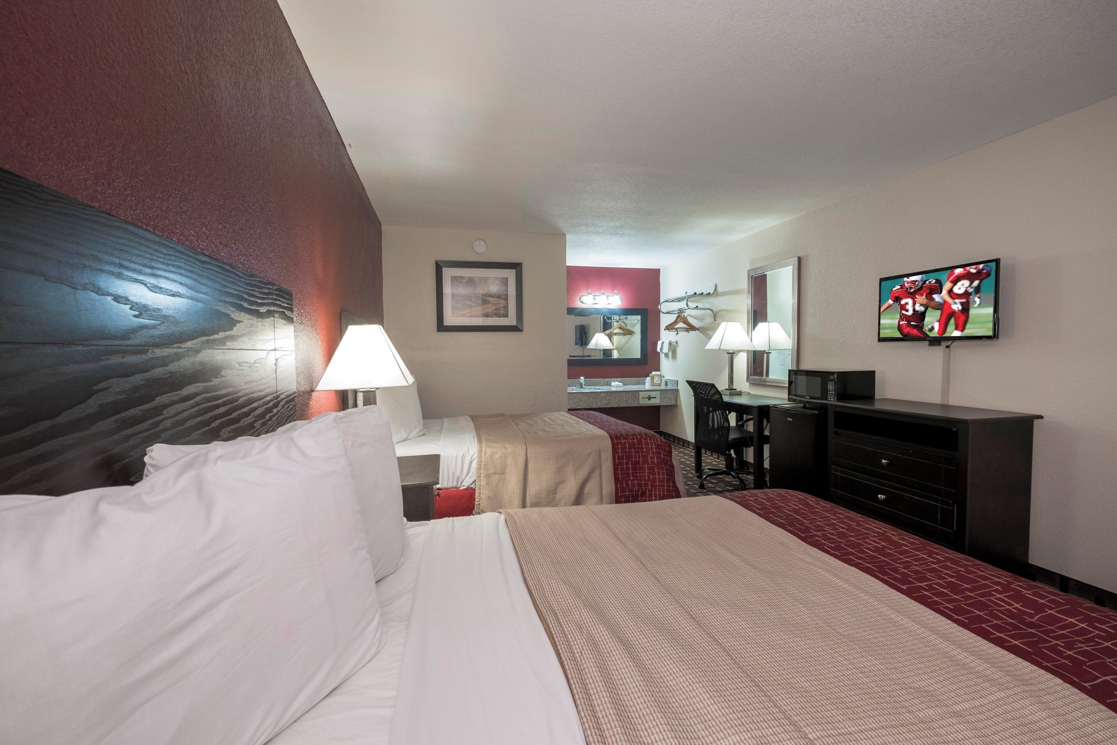 A hotel room with two beds, a desk, a mirror, a TV, and a red carpet.