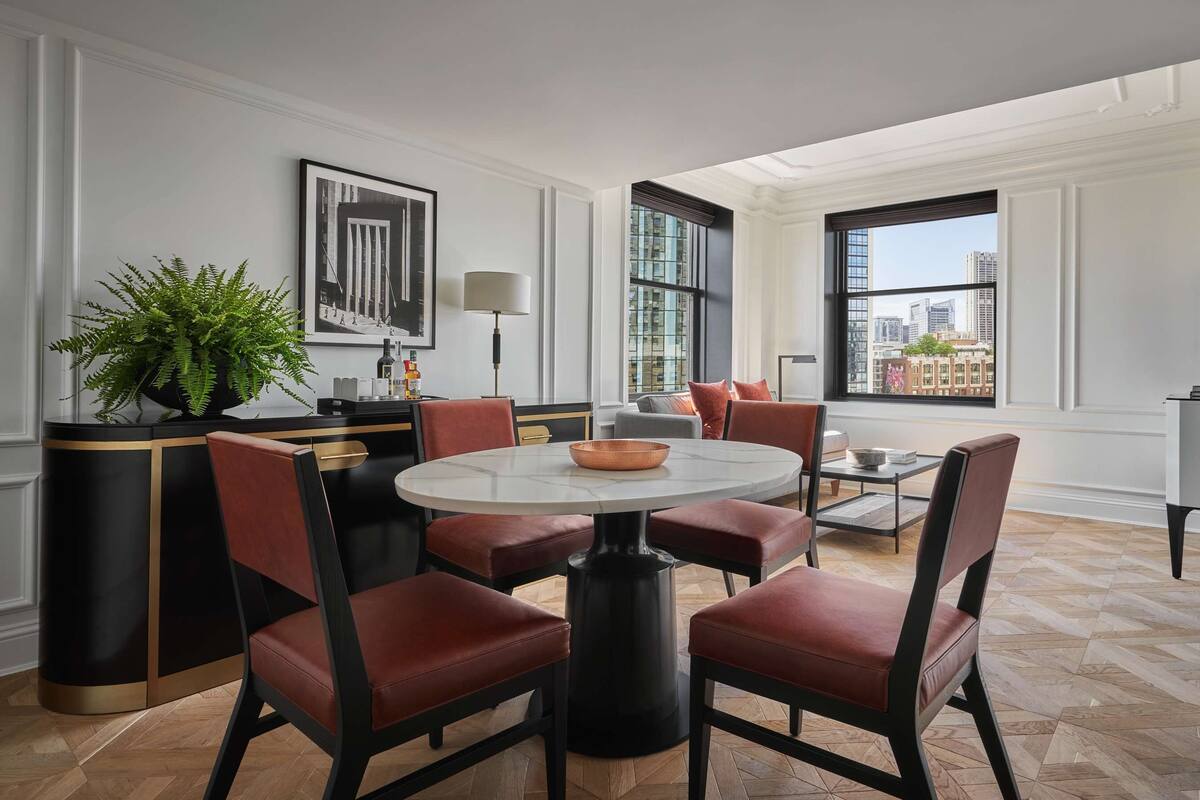 A modern living room with a round table, red chairs, a coffee table, a TV, and large windows with city views.