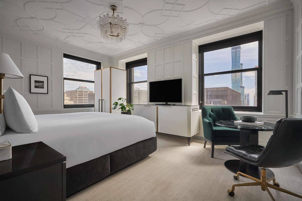 A modern hotel room with a large bed, a desk with a chair, a television, and large windows offering city views.
