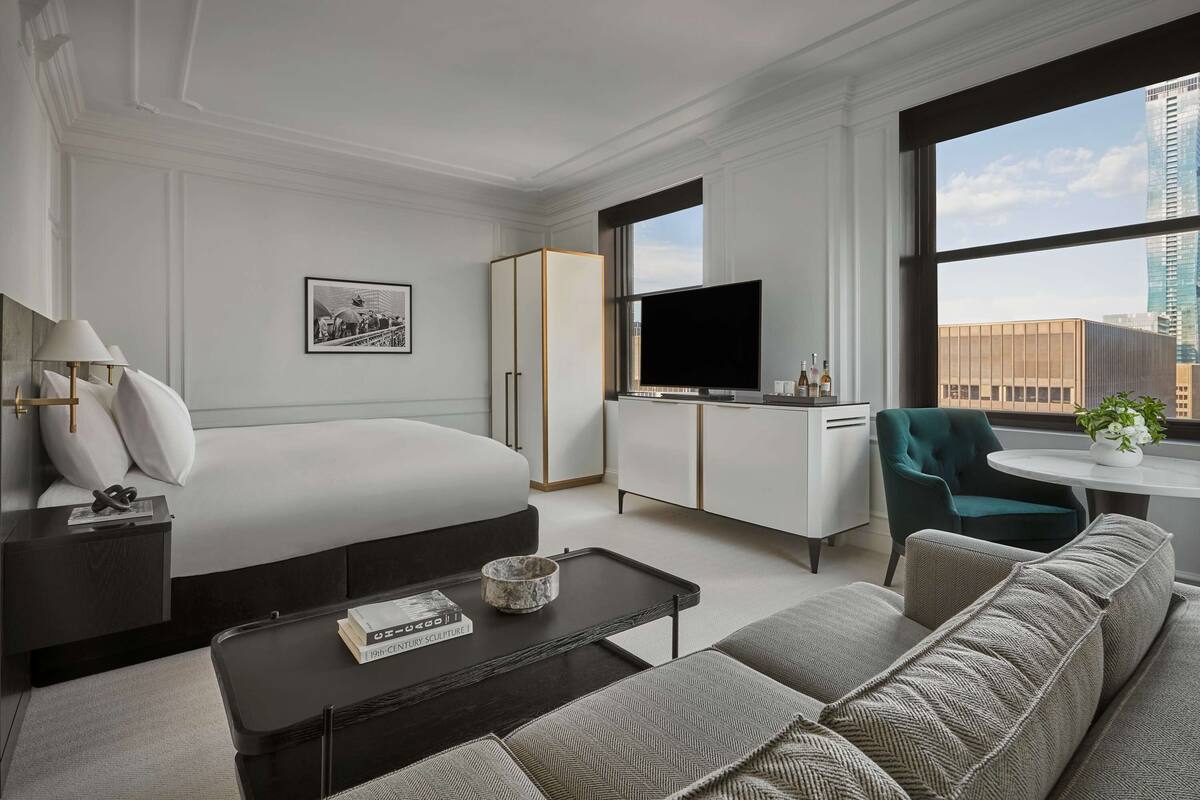 A modern hotel room with a bed, a sofa, a TV, a desk, and a window with a city view.