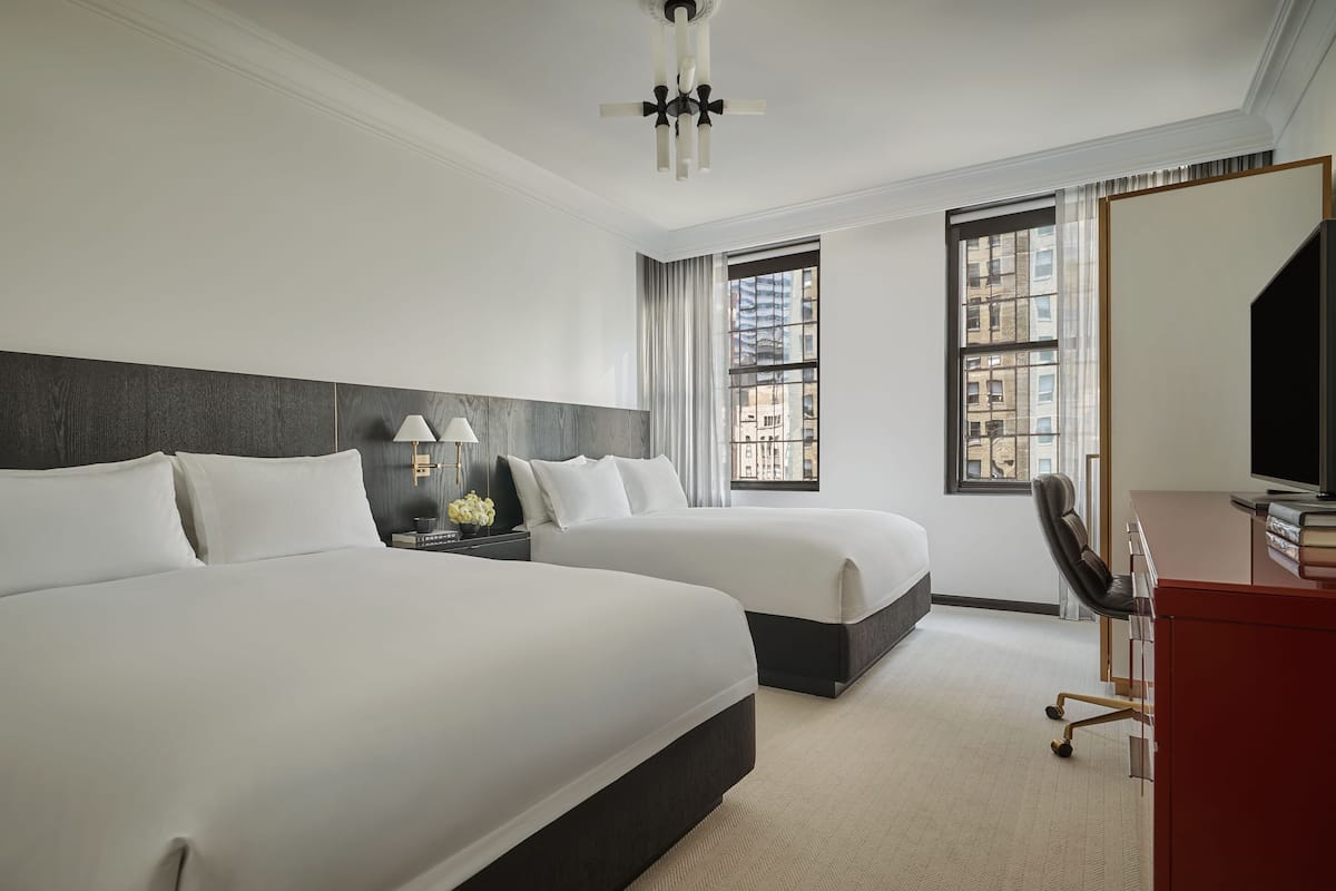A hotel room with two beds, a desk, a chair, and a large window with a city view.