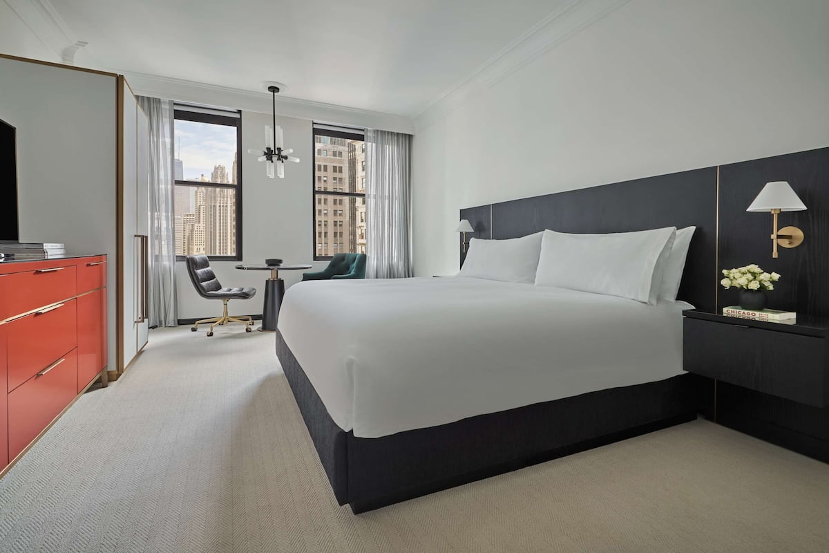 A hotel room with a large bed, a desk, a chair, a red dresser, and a window with a city view.