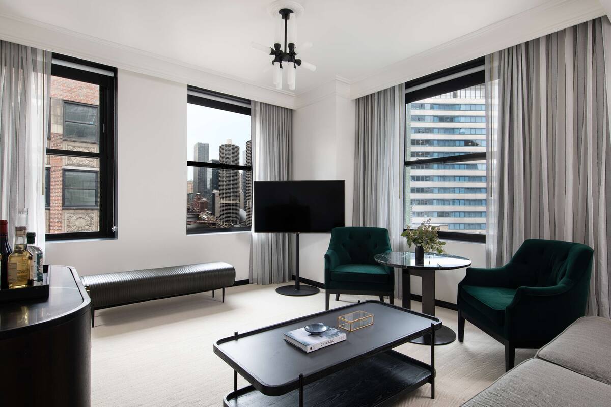A modern hotel room with a large window, a flat-screen TV, a green armchair, a sofa, and a small table with a book.