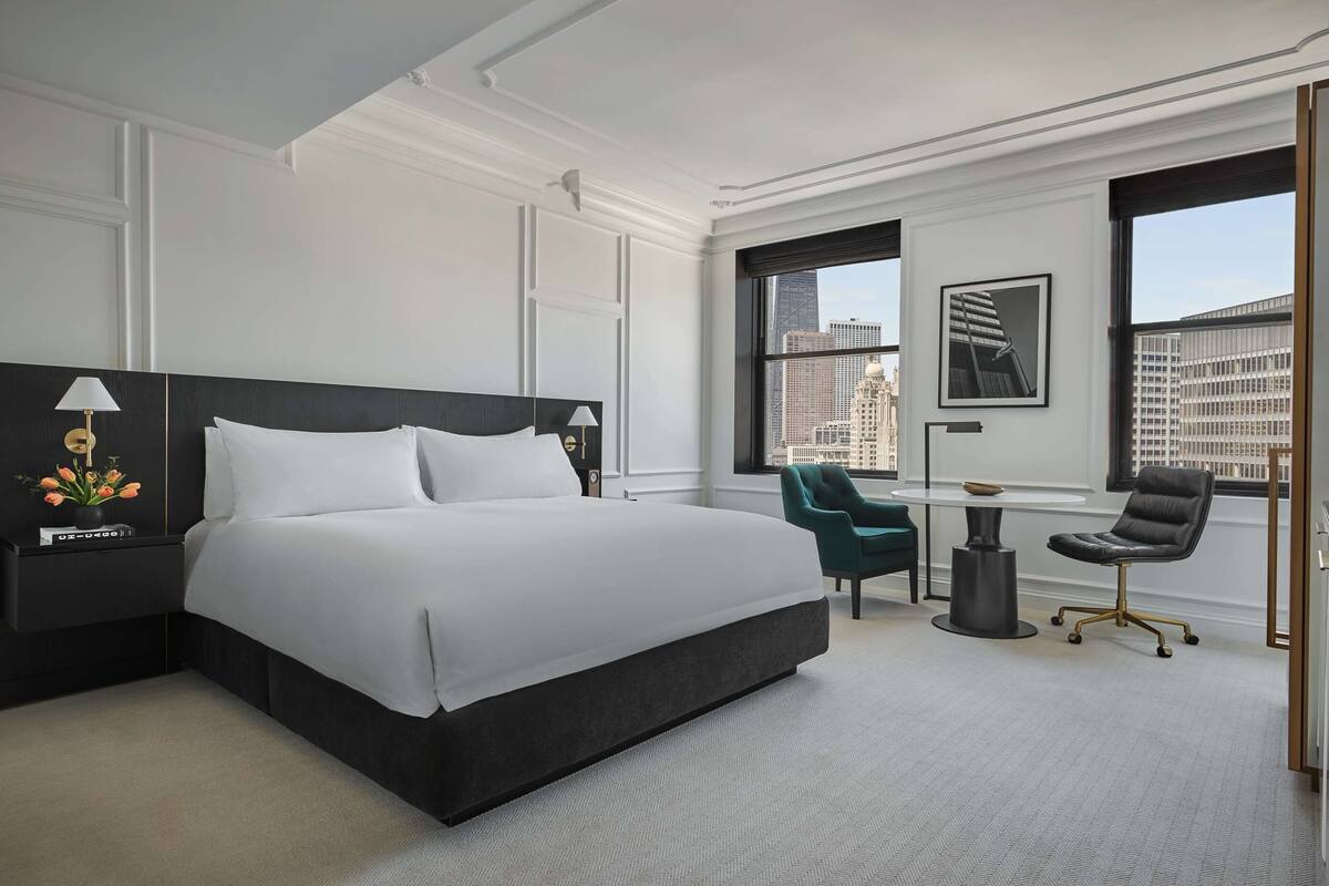 A modern hotel room with a large bed, a desk, a chair, and a view of the cityscape.