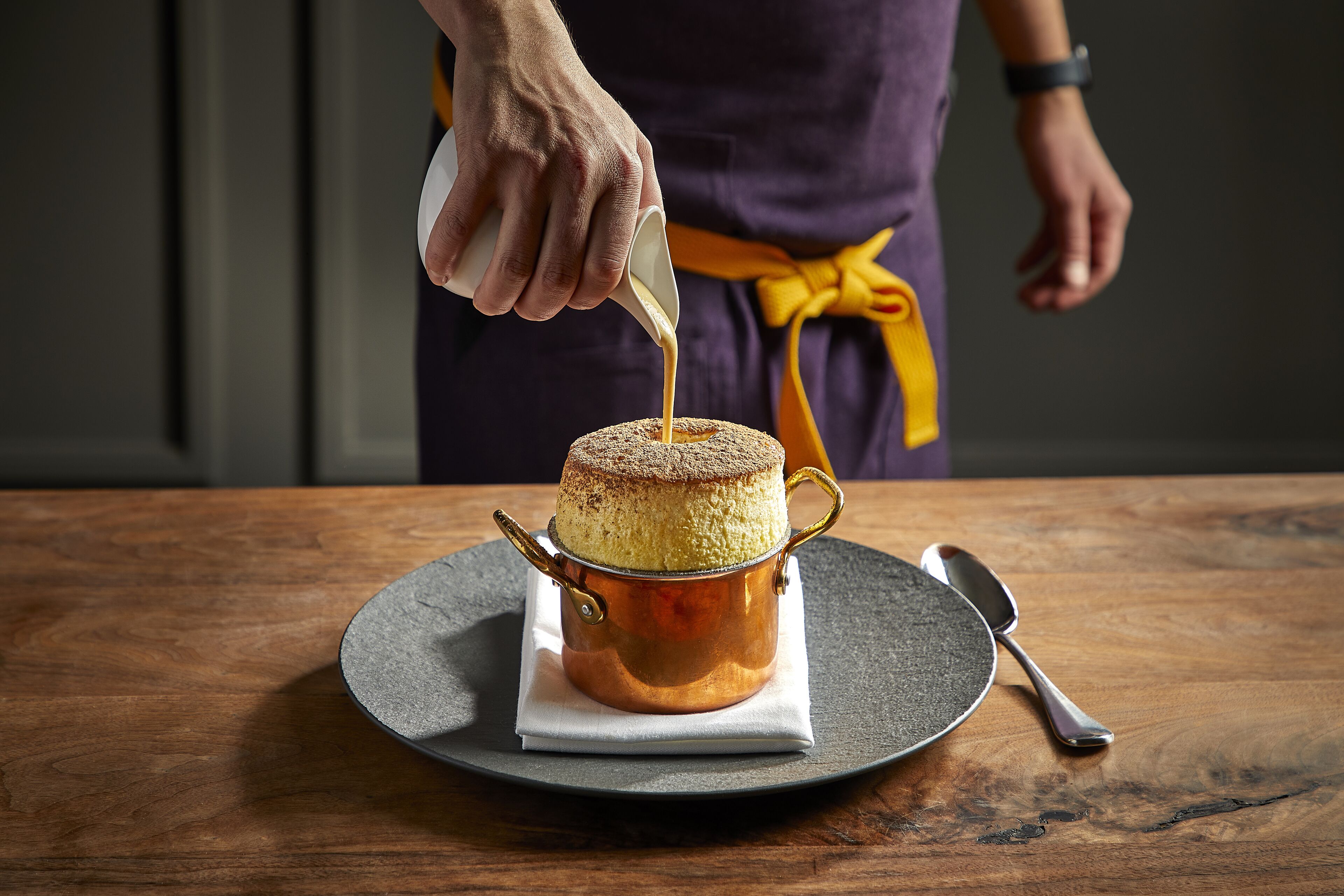 Un gâteau dans un récipient en cuivre, posé sur une table en bois, avec une cuillère et une serviette à proximité.