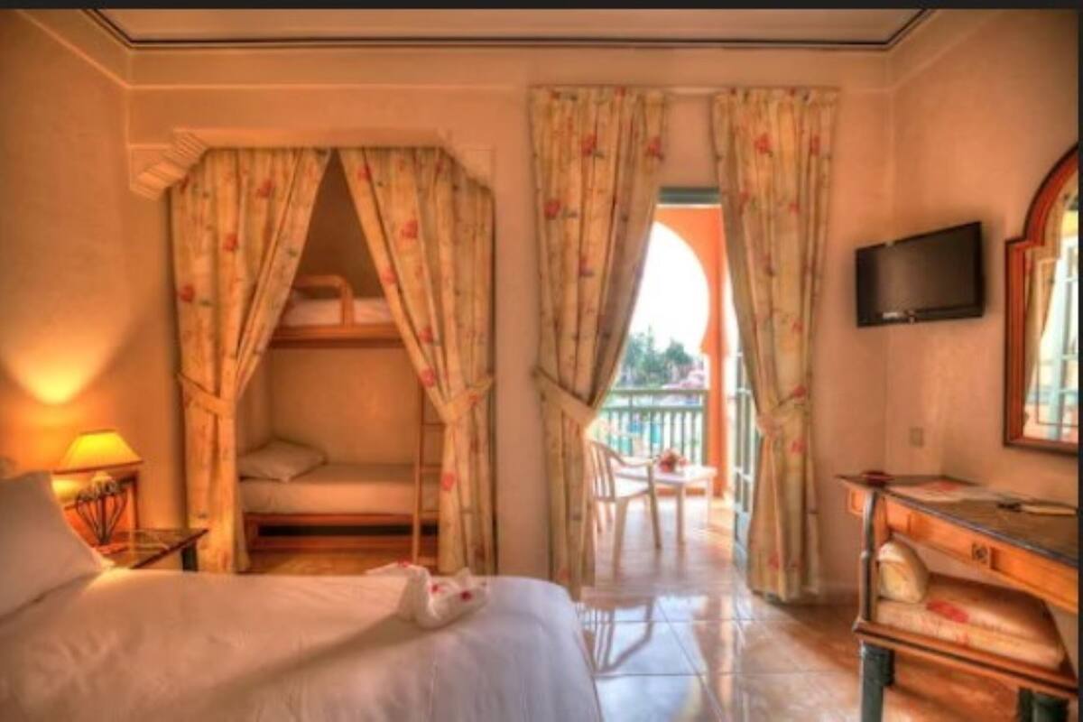 A bedroom with a bed, a bedside table, a lamp, a TV mounted on the wall, and a balcony with a chair and table.