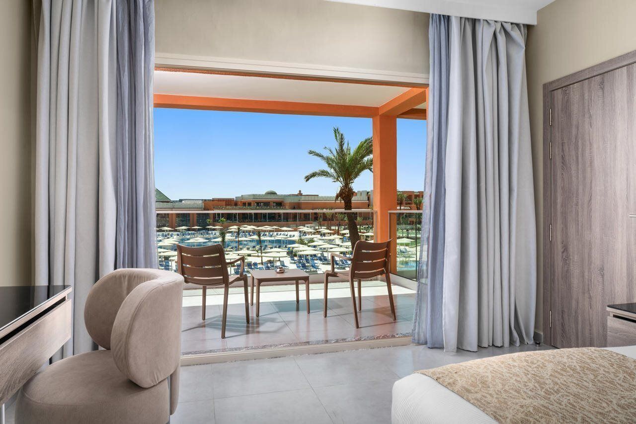 A hotel room with a balcony, a desk, a chair, and a view of a pool and resort buildings.