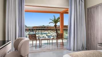 A hotel room with a balcony, a desk, a chair, and a view of a pool and resort buildings.