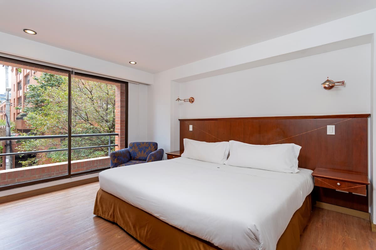 Camera d'albergo con un letto grande, un comodino, una sedia e una finestra con vista su edifici e alberi.