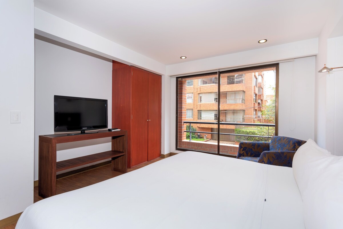 A bedroom with a bed, a TV on a wooden stand, a wardrobe, and a balcony with a view of buildings.