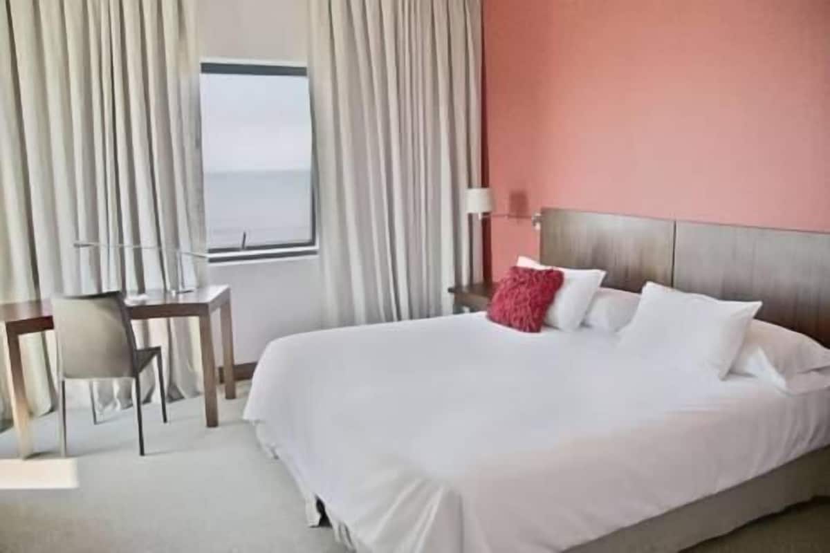 A hotel room with a large bed, a desk, and a chair. There is a window with curtains, a lamp, and a view of the sea.