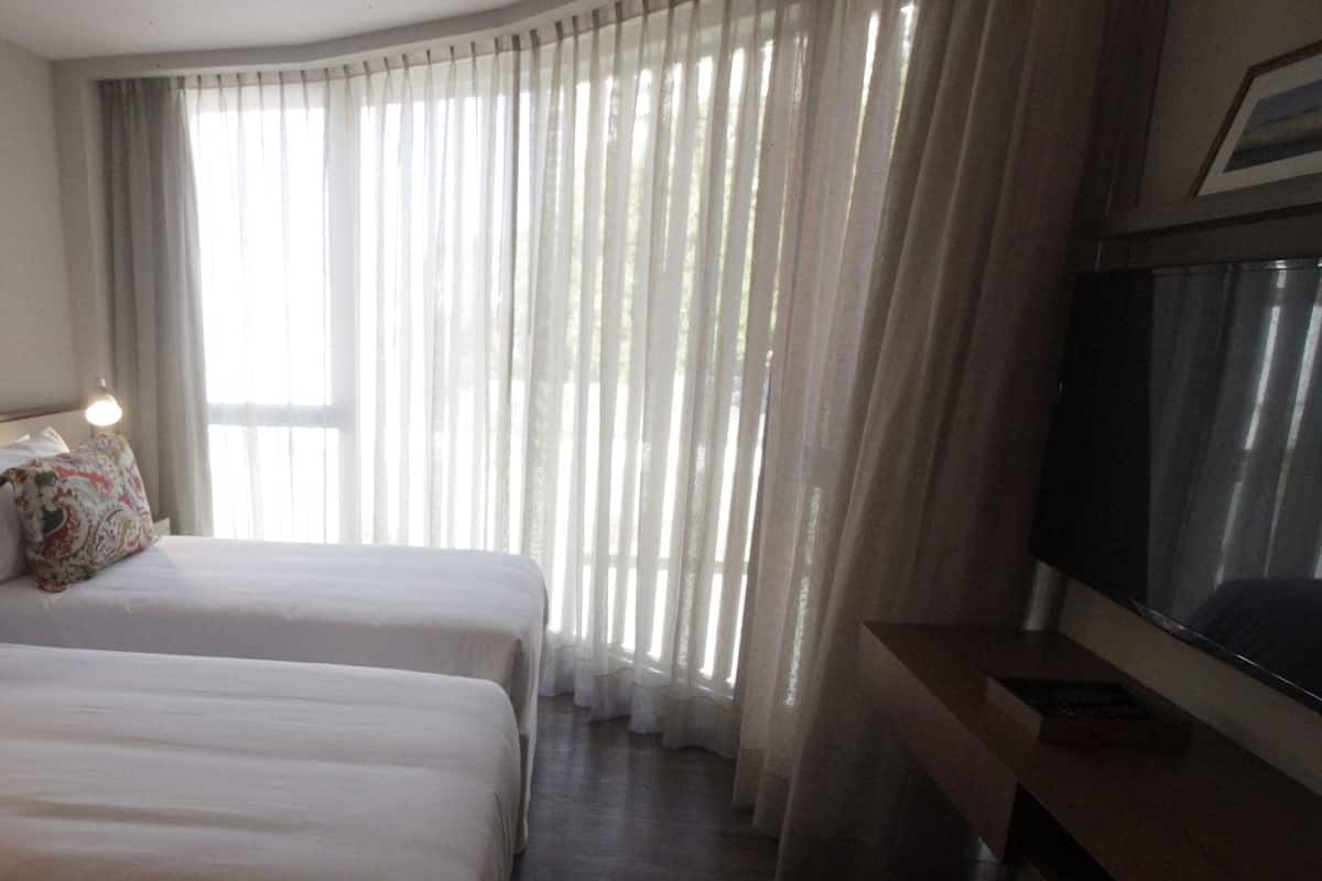 A hotel room with two beds, a TV, and a large window with sheer curtains.
