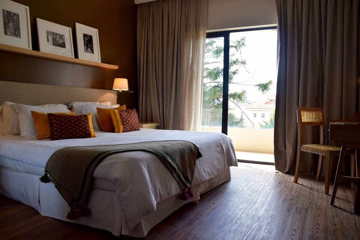 A hotel room with a large bed, a desk, and a chair. There is a window with curtains, and a wall with framed pictures.