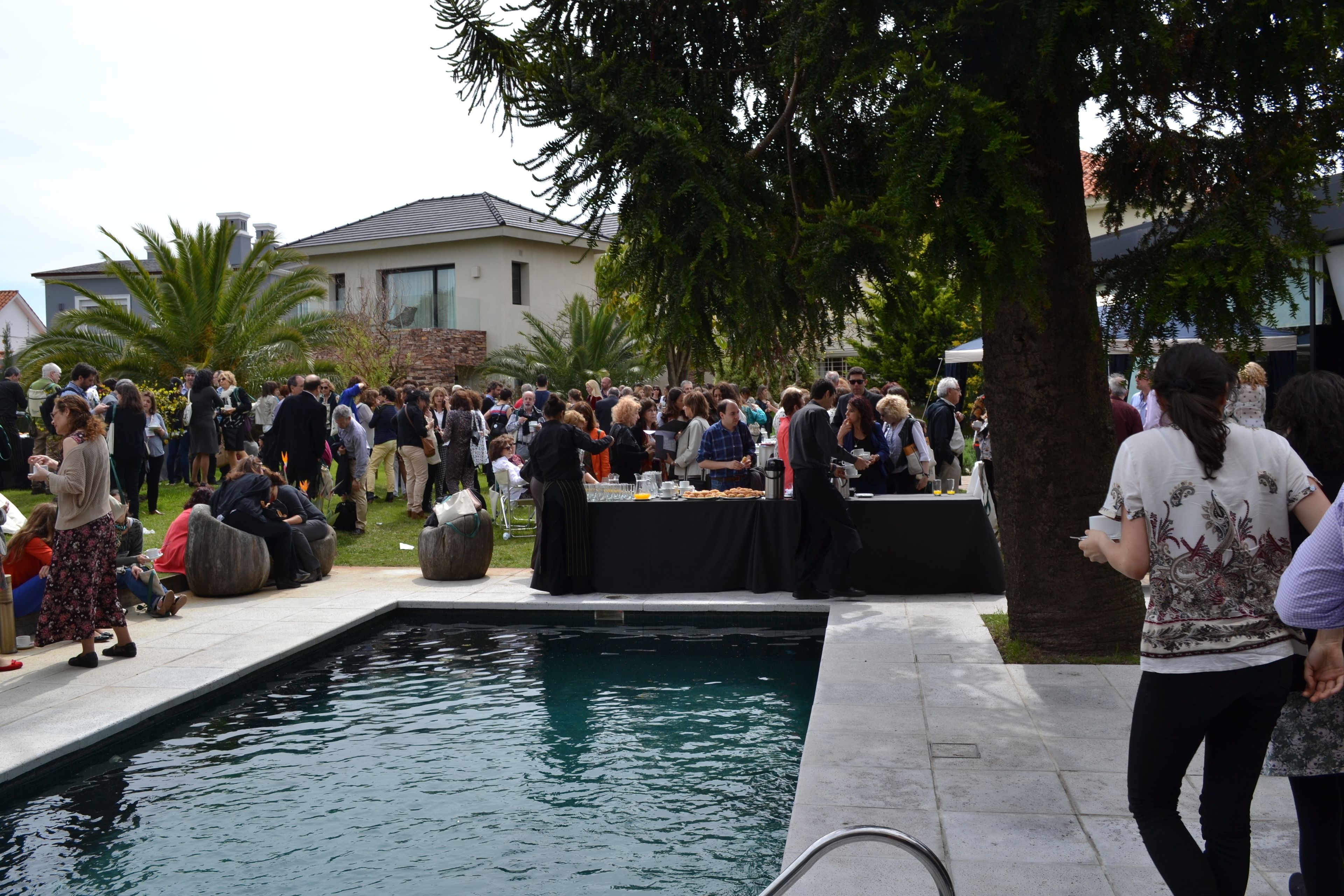 Um grupo de pessoas reunido junto a uma piscina, com uma mesa de buffet, e um edifício ao fundo.