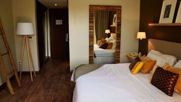 Habitación de hotel con una cama grande, una mesita de noche con lámpara, suelo de madera y una puerta que da a otra habitación.