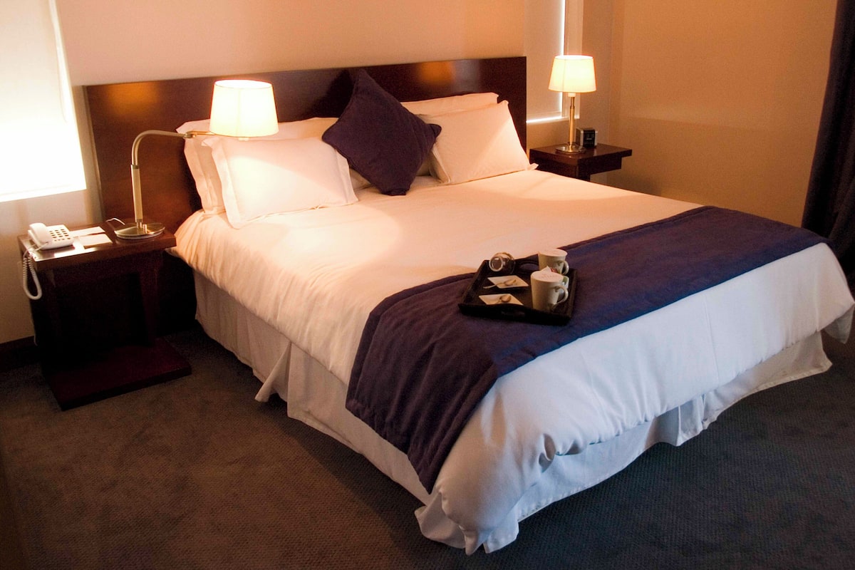 A hotel room with a large bed, two bedside tables with lamps, a telephone, and a tray with a cup and a remote control.