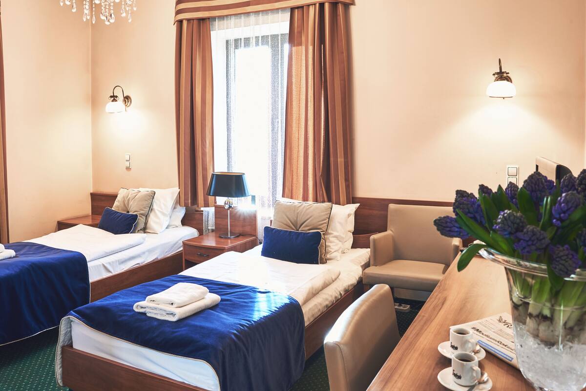 A hotel room with two single beds, a desk with a chair, a small table with two cups, and a vase with flowers.