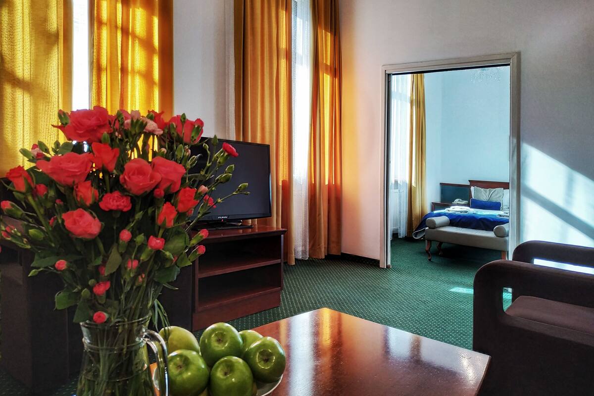 A hotel room with a bed, a TV, a table with a bowl of apples, and a vase of red roses.