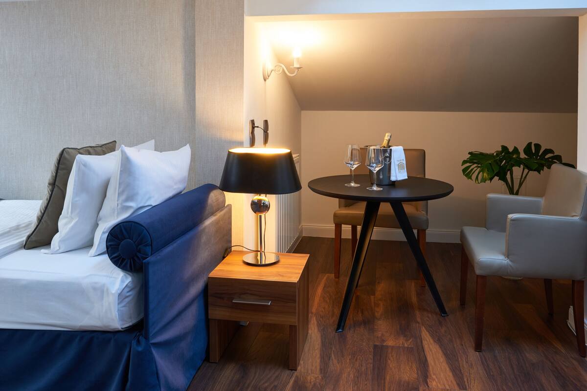 A hotel room with a bed, a bedside table with a lamp, a small table with a chair, and a potted plant.