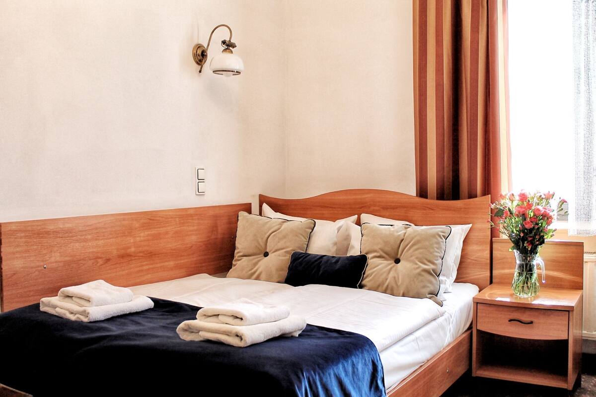 A neatly made bed with a dark blue bedspread and white linens, a wooden headboard, and a bedside table with a vase of flowers.