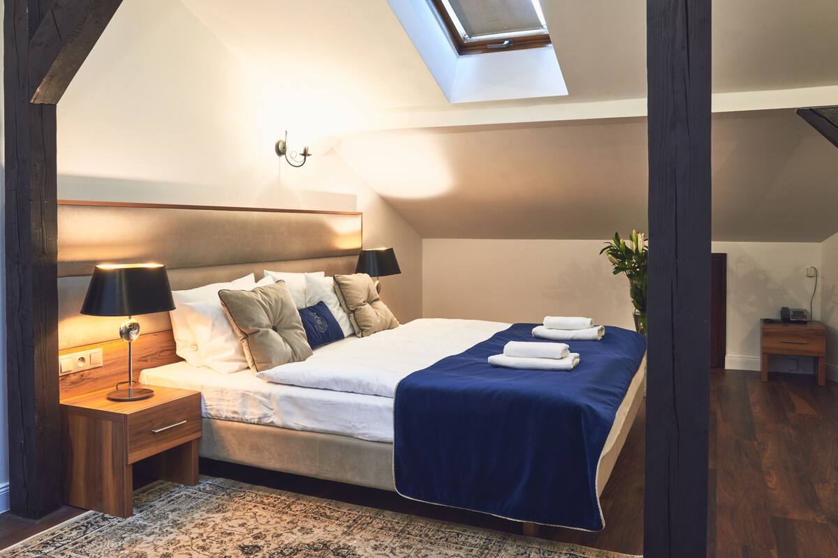 A bedroom with a skylight, a bed with a blue bedspread, a nightstand with a lamp, and a wooden floor.