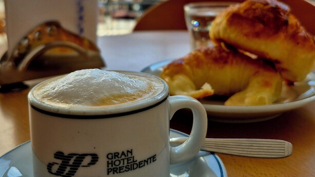 A cup of coffee with a logo, a croissant, and a plate with a pastry on a table. - Gran Hotel Presidente (Salta)