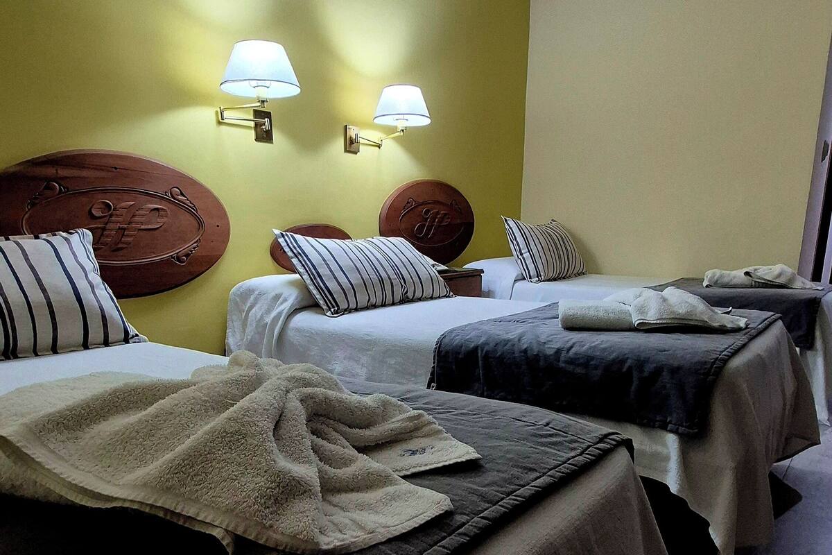 Ein Hotelzimmer mit drei Betten, jeweils mit Kopfstütze und Kissen. An beiden Seiten der Betten befinden sich Wandlampen.