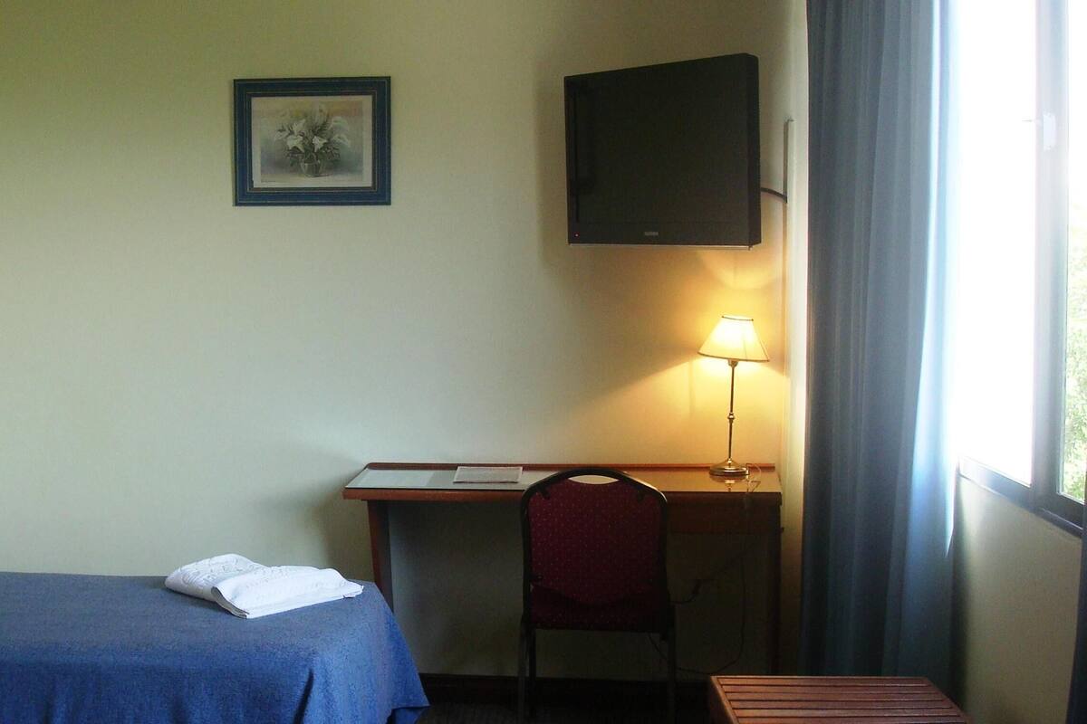 Ein Hotelzimmer mit einem Einzelbett, einem Schreibtisch mit Stuhl, einem an der Wand befestigten Fernseher und einem Fenster mit Vorhängen.