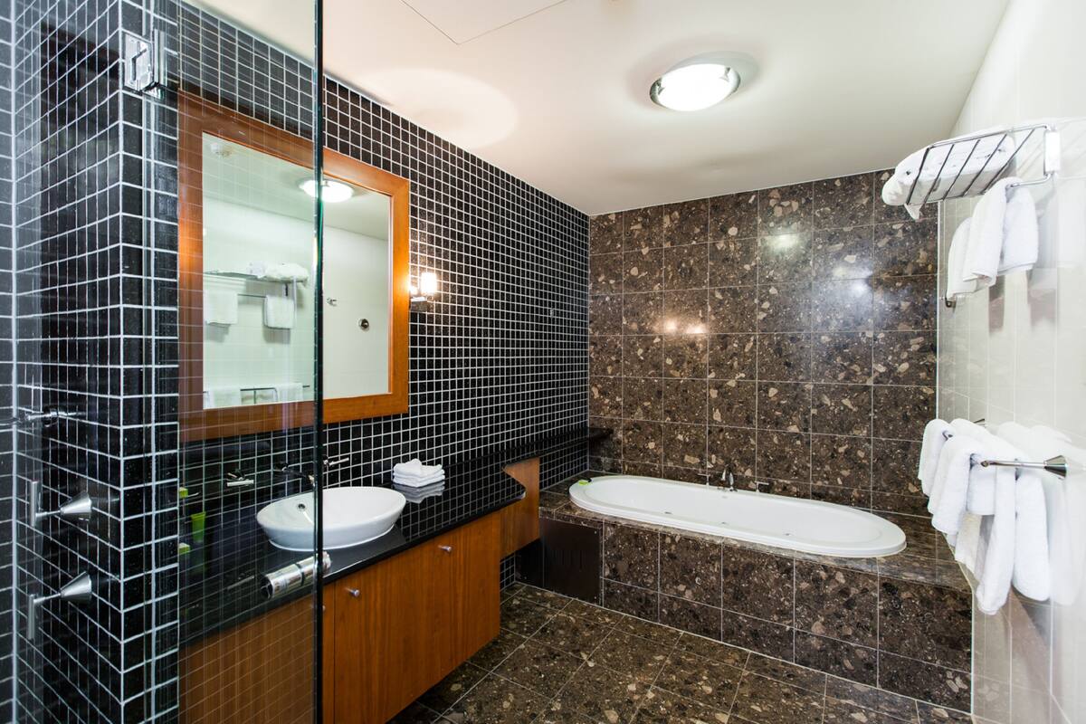 Un baño moderno con pared de azulejos negros, tocador de madera, lavabo blanco, espejo, bañera y mampara de vidrio.