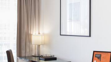 Une chambre d’hôtel équipée d’un bureau, d’une lampe, d’un tableau encadré au mur et d’une fenêtre avec des rideaux.