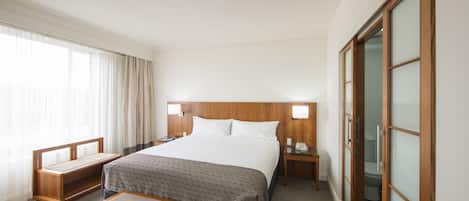 Habitación de hotel con una cama grande, mesitas de noche, un banco de madera, una pequeña mesa y un armario con puerta corredera.