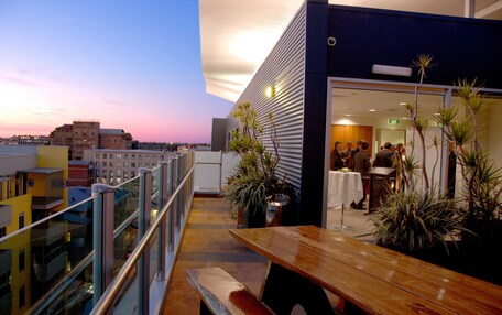 A rooftop terrace with a wooden table and benches, potted plants, and a view of the cityscape.