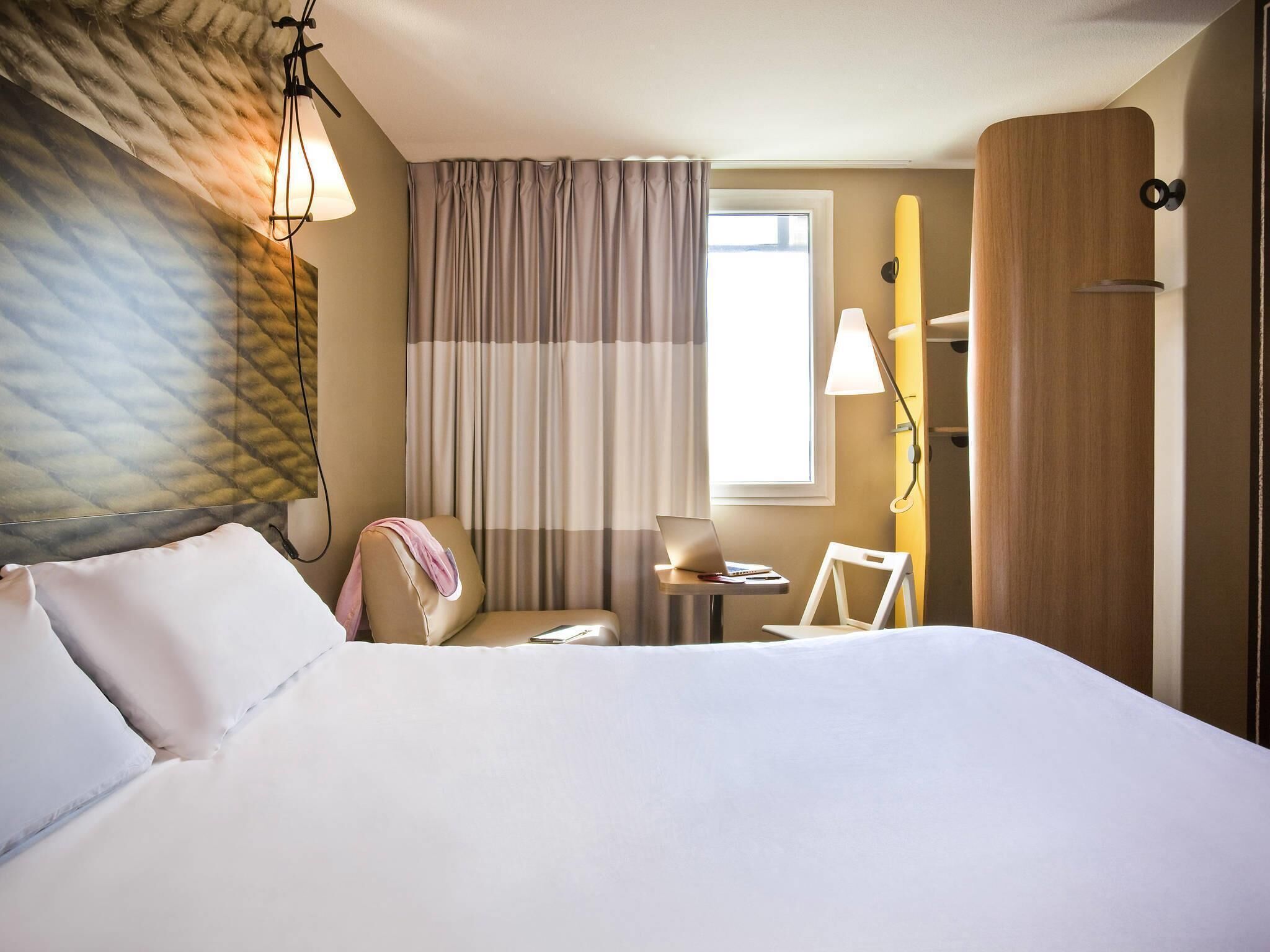 Ein Hotelzimmer mit einem Bett, einem Schreibtisch mit Laptop, einem Stuhl und einem fenster mit Vorhängen.