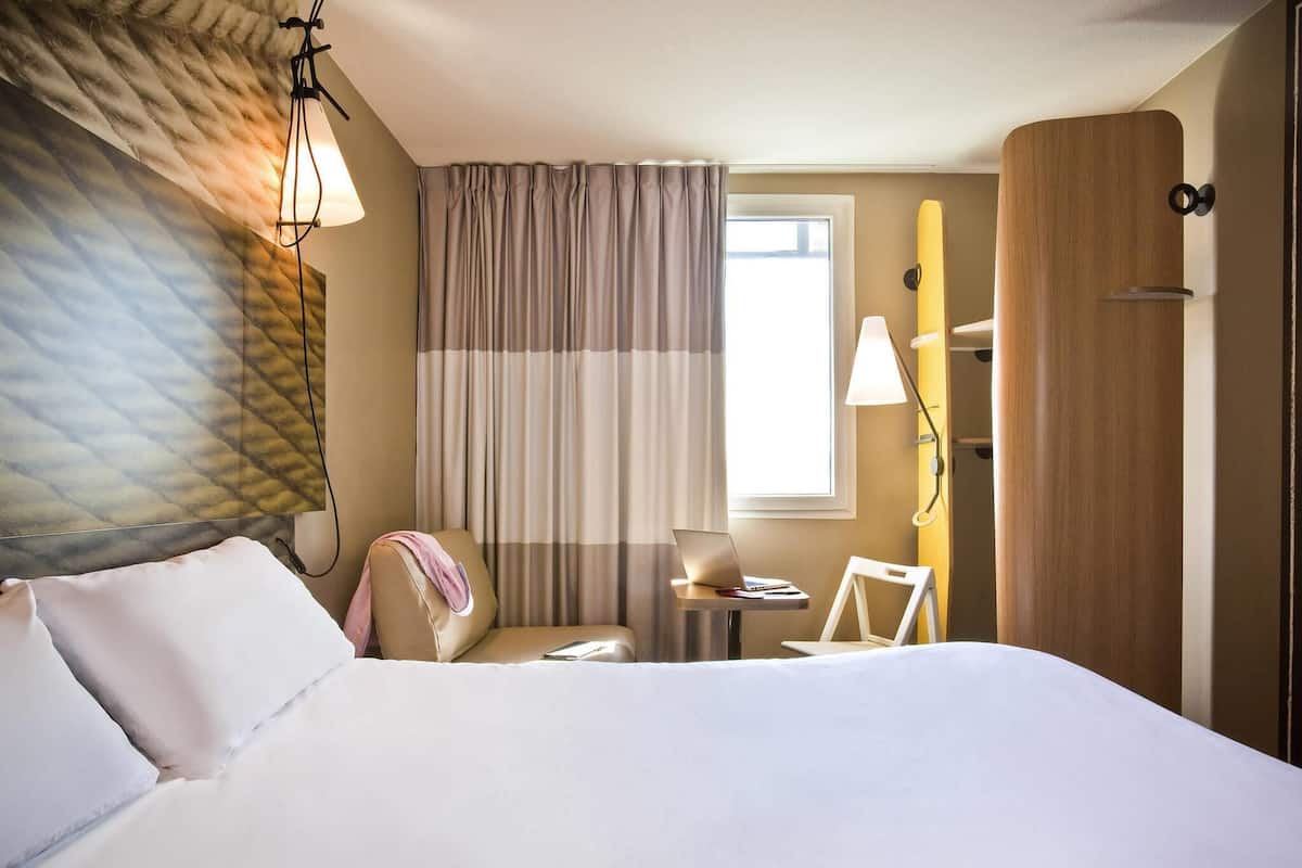 Ein Hotelzimmer mit einem Bett, einem Schreibtisch mit Laptop, einem Stuhl und einem fenster mit Vorhängen.
