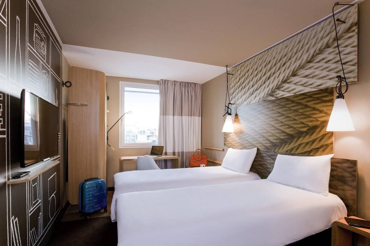 Hotelkamer met twee bedden, een televisie, een bureau met een laptop, een koffer en een raam met gordijnen.