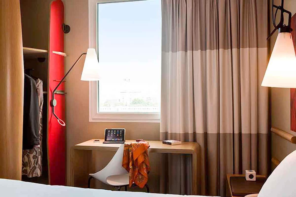 Een hotelkamer met een bed, een bureau met een laptop, een stoel, een rode surfplank, een raam met gordijnen en een lamp.