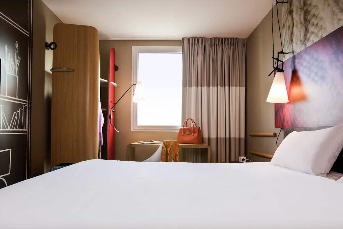 Een hotelkamer met een bed, nachtkastje, lamp, raam met gordijnen en een kunstwerk aan de muur.