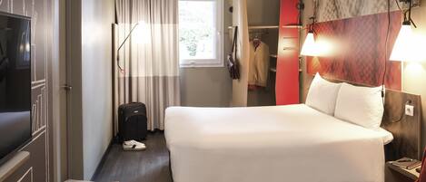 A hotel room with a bed, a desk with a magazine and glasses, a suitcase, a coat rack with clothes, and a TV.