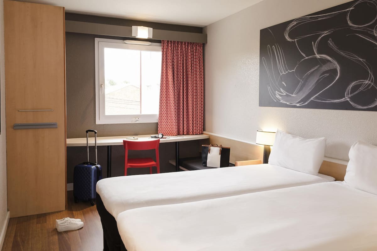 Ein Hotelzimmer mit Bett, Schreibtisch, Stuhl, Koffer und einem Fenster mit Vorhängen.