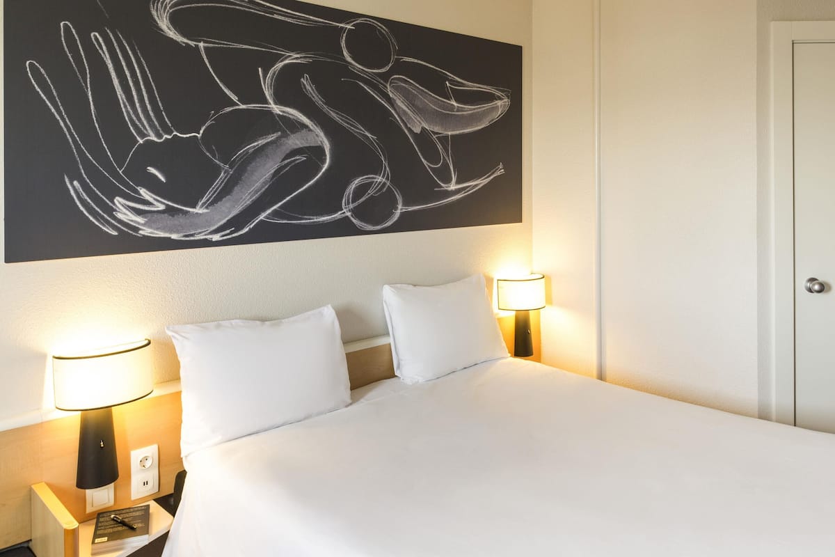 Ett hotellrum med en stor, abstrakt målning på väggen, två sänglampor, en säng med vita sängkläder och ett nattduksbord med en bok och en lampa.